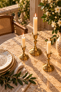 Three brass candlesticks holding lit white taper candles arranged on a sunlit stone table, with plates, a woven chair, and a floral arrangement in the background.